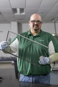 man standing in a lab setting wearing a green polo shirt that says Quanex, holding a glass unit with no frame