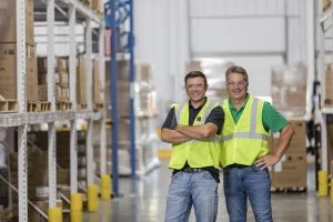 two men standing in a warehouse setting with green safety vests